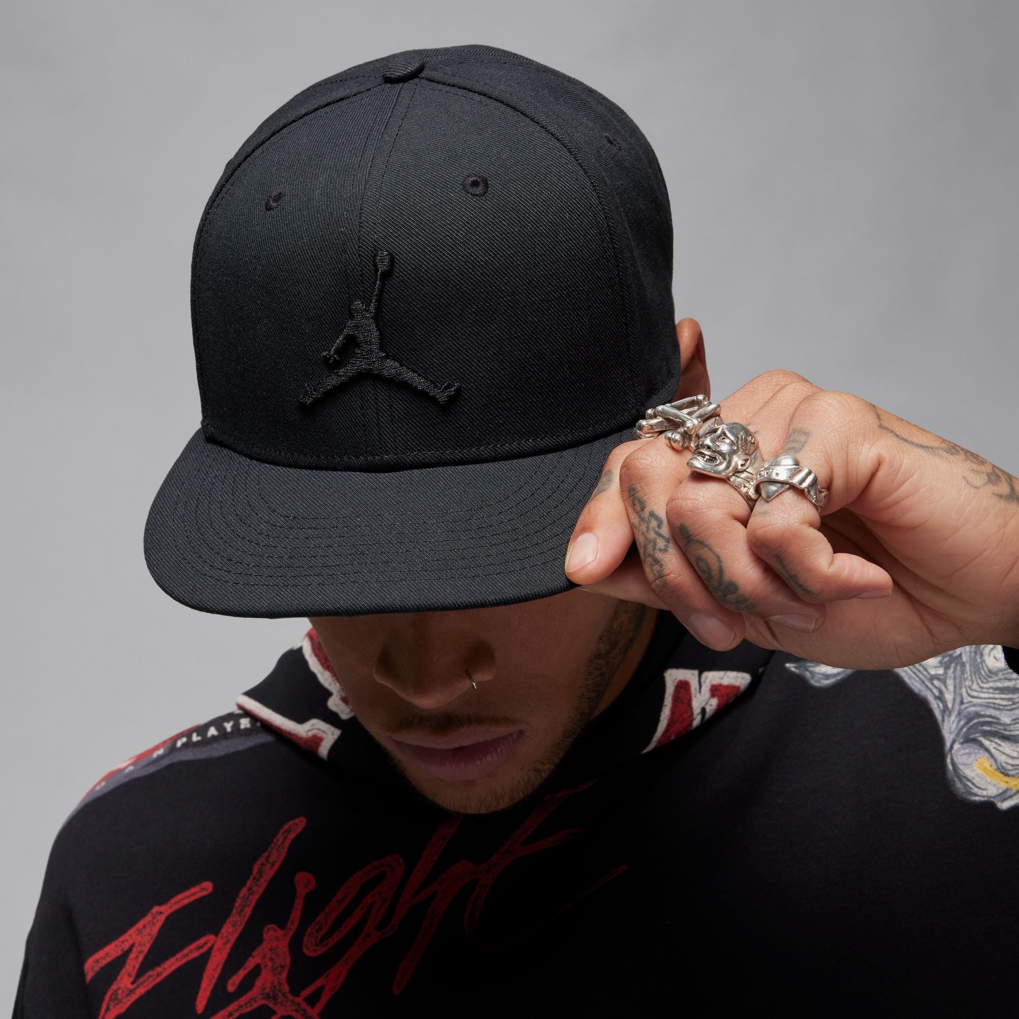Person wearing a black cap with a logo, holding it to their face against a gray background