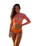Woman in a colorful floral long-sleeve top and orange bikini bottom standing on a beach.