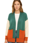 A woman wearing a color-blocked knit cardigan with a teal green body, cream sleeves, and orange pocket panels, styled over a white t-shirt and jeans against a plain white background.