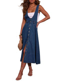 A woman models a blue denim overall midi dress with a button-front closure and a high side slit, worn over a white tank top. She is holding a woven clutch and wearing strappy high-heeled sandals, standing against a plain beige wall.