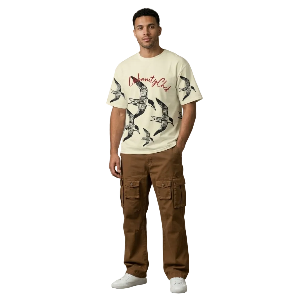 Man wearing a beige t-shirt with graphic design and brown cargo pants against a gray concrete wall.