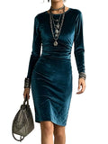 A woman wearing a long-sleeve teal velvet knee-length dress, accessorized with layered silver necklaces, bracelets, and a gray chain shoulder bag, standing against a textured gray wall.
