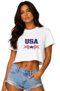 A woman modeling a white cropped t-shirt with a red, white, and blue 'USA' graphic on the chest, paired with light-wash denim shorts, against a neutral gray studio background.