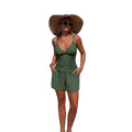 A woman stands on a sandy beach wearing an olive green two-piece swimsuit with a wide-brimmed straw hat and sunglasses, with the ocean and blue sky in the background.
