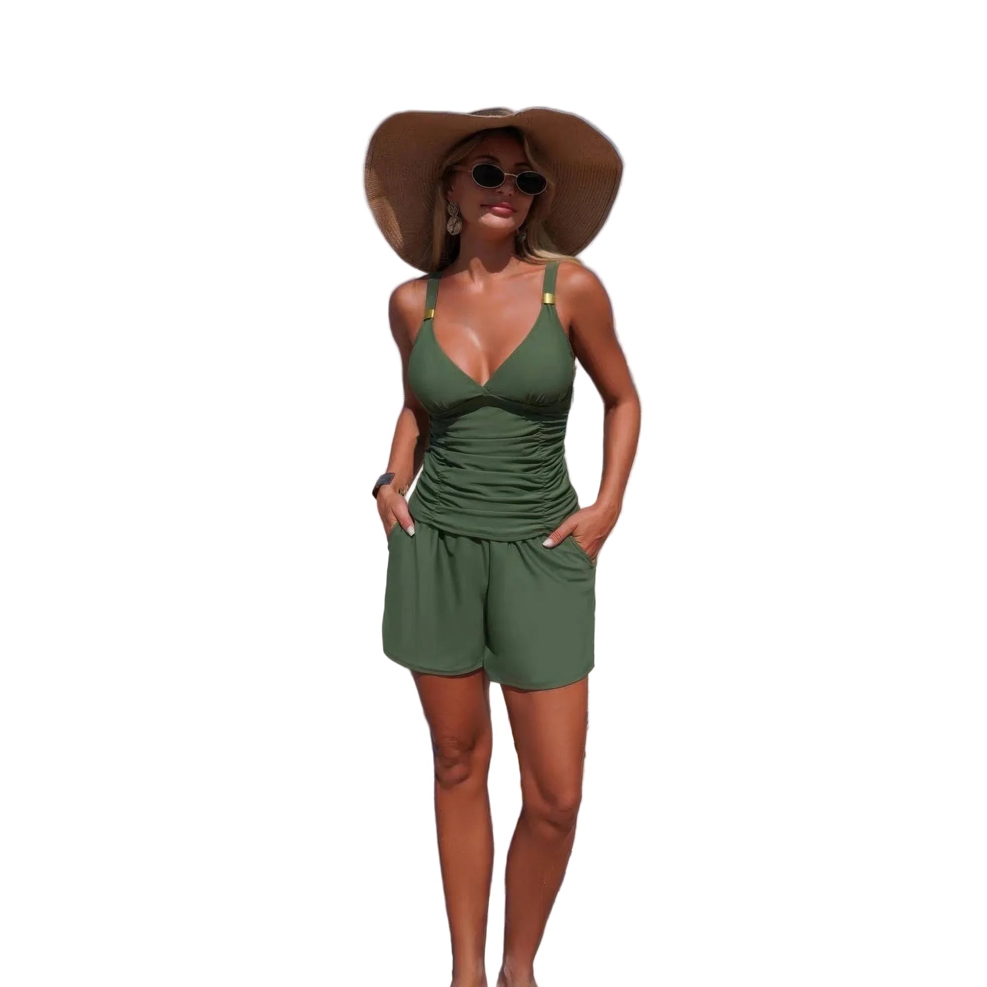 A woman stands on a sandy beach wearing an olive green two-piece swimsuit with a wide-brimmed straw hat and sunglasses, with the ocean and blue sky in the background.