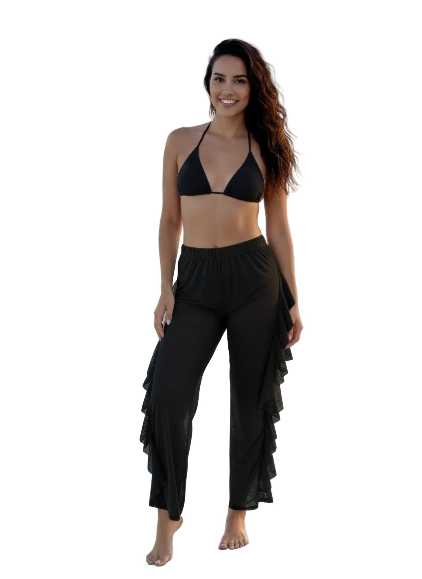 Woman in black bikini top and ruffled pants standing on a beach with ocean and palm trees in the background.