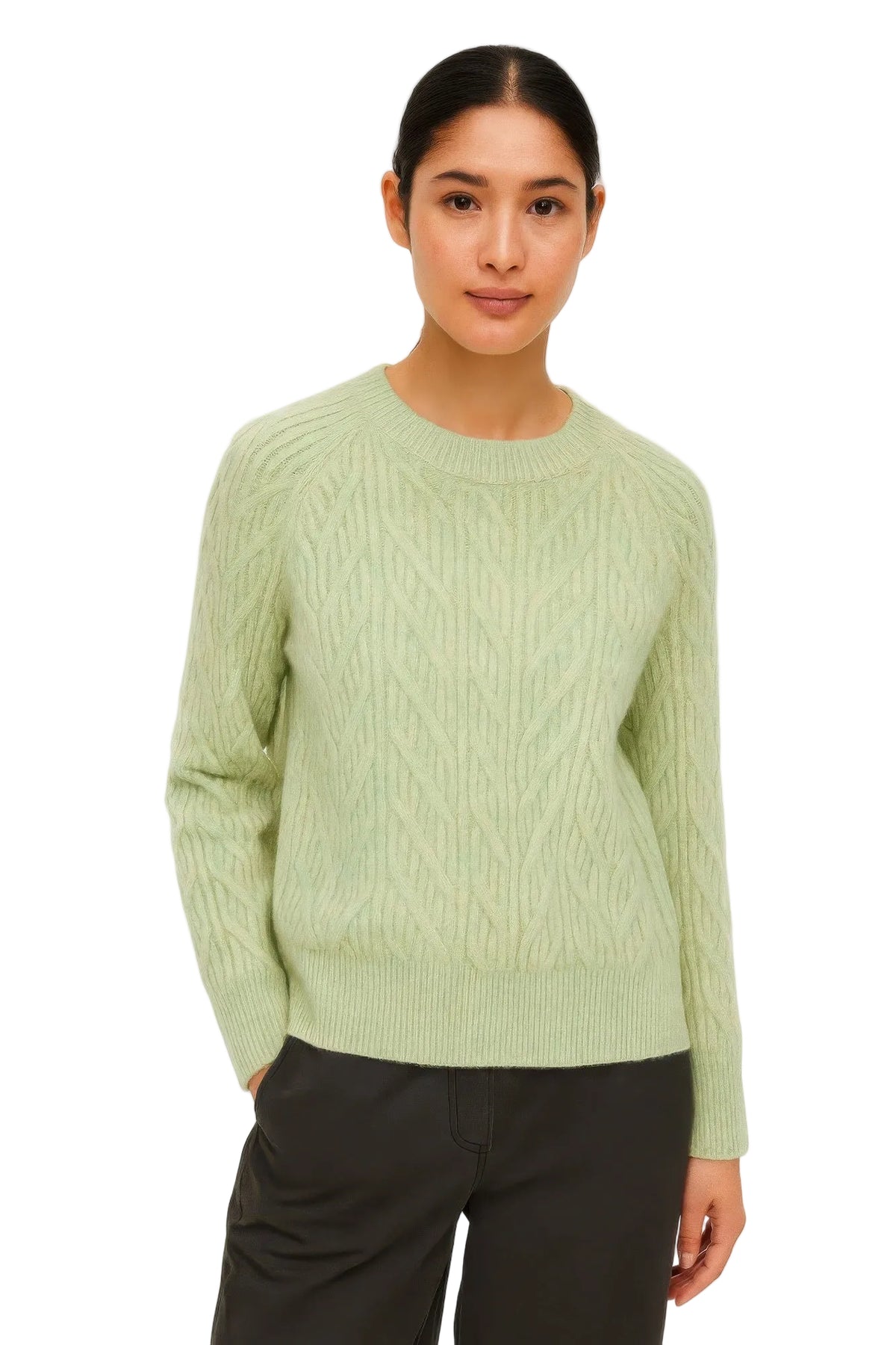 A woman modeling a light sage green cable-knit crewneck sweater with dark charcoal pants, shot against a plain white studio background.