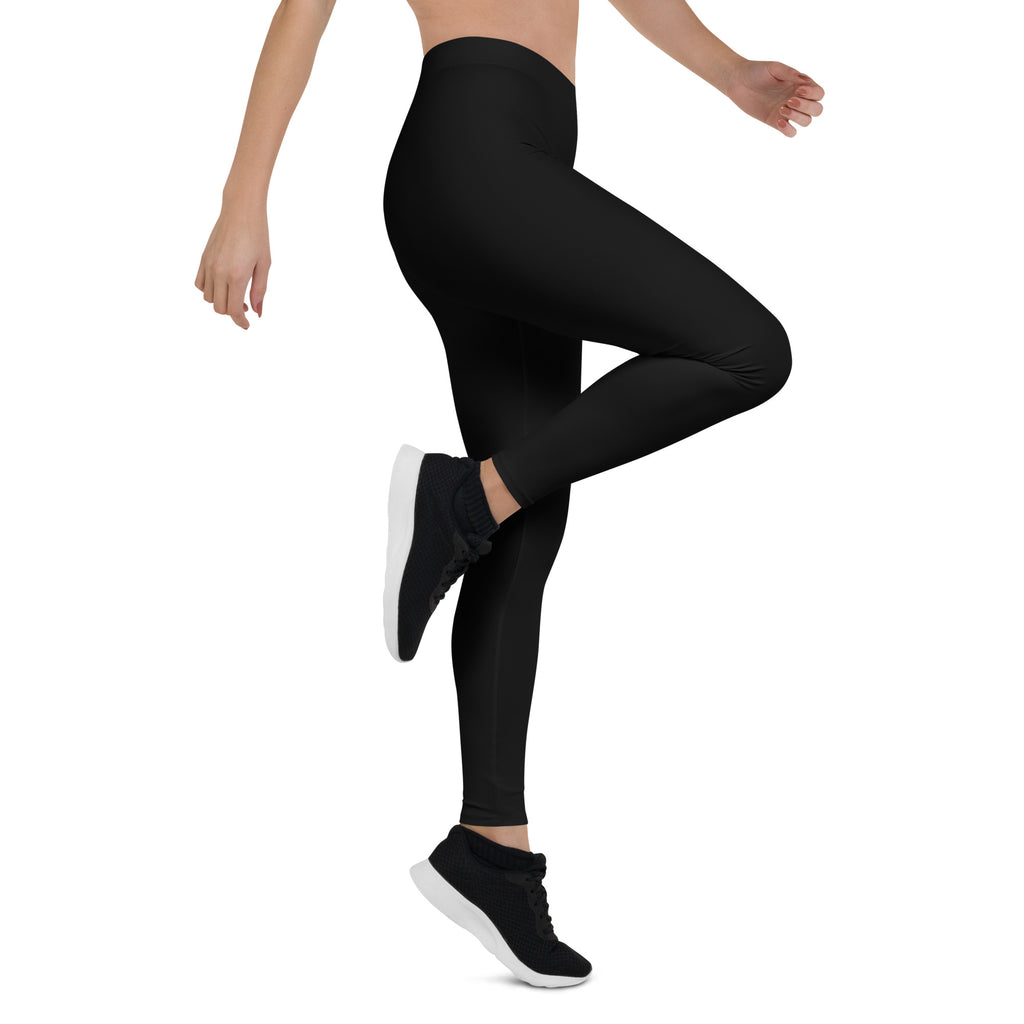 A woman posing in mid-rise black leggings and a pair of sneakers. The leggings are form-fitting and appear to be made of a stretchy fabric.